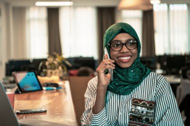 Gülümseyen genç bir kadın akıllı telefonda konuşur dizüstü bilgisayarda çalışır uzaktan düşünür. Modern cihazlar, cep telefonu ve bilgisayar kullanan mutlu bir Afrikalı Amerikalı müşteri. Yüksek kalite
