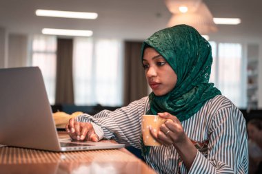Afrikalı Müslüman iş kadını yeşil tesettüre bürünmüş çay içerken aynı zamanda modern açık planlama ofisindeki rahatlama alanında dizüstü bilgisayarda çalışıyor..