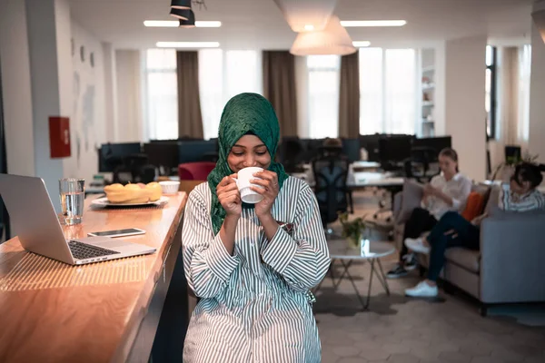 Afrikaanse moslim zakenvrouw het dragen van een groene hijab drinken van thee tijdens het werken op laptop computer in ontspanningsruimte bij moderne open plan startup office. — Stockfoto