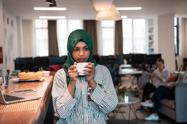 Afrikalı Müslüman iş kadını yeşil tesettüre bürünmüş çay içerken aynı zamanda modern açık planlama ofisindeki rahatlama alanında dizüstü bilgisayarda çalışıyor.. — Stok fotoğraf