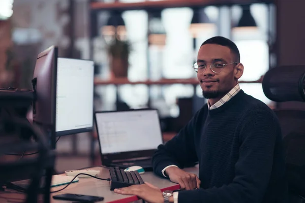 Молодой афроамериканец в очках сидит в современном офисе и работает за компьютером — стоковое фото