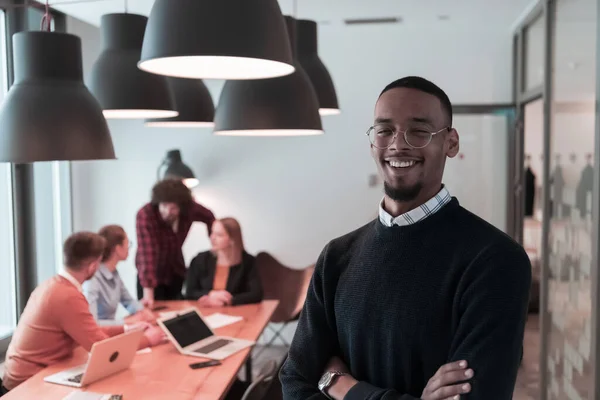 Portrét šťastného afroamerického tisíciletého majitele mužského byznysu v moderní kanceláři. Podnikatel nosí brýle, usmívá se a dívá se do kamery. Rušný různorodý tým pracující v pozadí. Koncept vedení — Stock fotografie