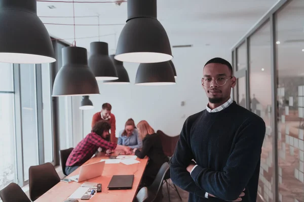 Portrait de heureux Afro Américain millénial propriétaire d'entreprise masculine dans le bureau moderne. Homme d'affaires portant des lunettes, souriant et regardant la caméra. Une équipe diversifiée et occupée qui travaille en arrière-plan. Concept de leadership — Photo