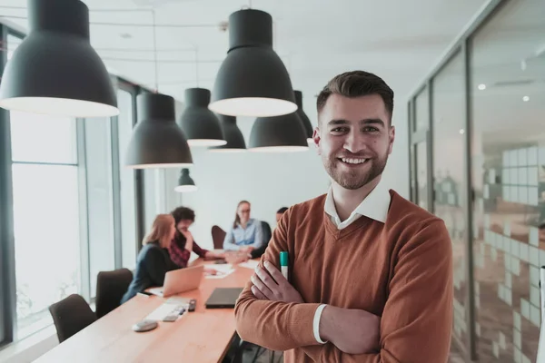 Egy boldog Y-generációs férfi üzletember portréja a modern irodából. Egy üzletember szemüveget visel, mosolyog és a kamerába néz. Elfoglalt, változatos csapat dolgozik a háttérben. Vezetői koncepció. Fejlövés. — Stock Fotó
