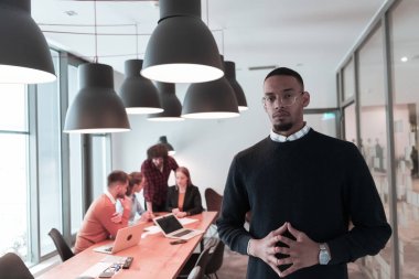 Modern ofisteki mutlu Afro-Amerikan erkek iş adamının portresi. İş adamı gözlük takıyor, gülümsüyor ve kameraya bakıyor. Arka planda çalışan çeşitli ekipler. Liderlik kavramı