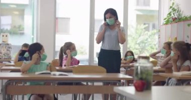 Sınıfta çalışan, sınıfta çalışan, öğretmenlerin açıklamalarını dinleyen ve yazan çok ırklı maskeler takan bir grup çocuk.