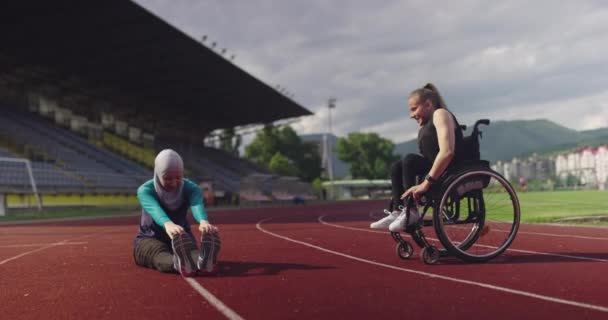 Kobieta z niepełnosprawnością rozgrzewa się na wózku inwalidzkim na torze treningowym lekkoatletycznym z muzułmanką ubraną w strój sportowy hidżab — Wideo stockowe