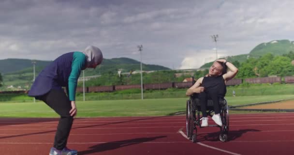 Žena s postižením zahřívání na invalidním vozíku na atletické tréninkové dráze s muslimskou ženou na sobě hidžáb sportovní oblečení — Stock video