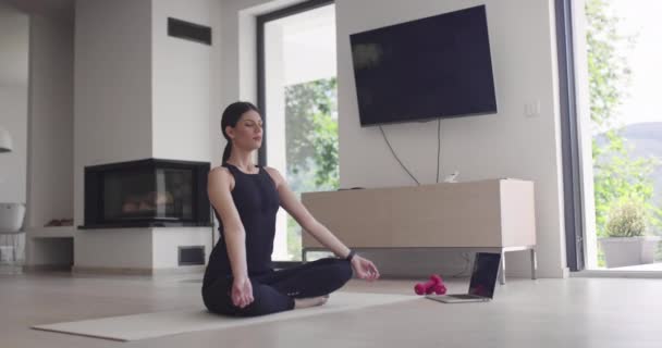 Mujer en ropa deportiva haciendo ejercicios de estiramiento mientras ve clases de entrenamiento de yoga en la computadora portátil en línea. Chica sana haciendo ejercicio en la sala de estar con sofá sofá en el fondo.. — Vídeos de Stock