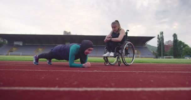 Una mujer con discapacidades calentándose en silla de ruedas en pista de entrenamiento de atletismo con una mujer musulmana usando ropa deportiva de hijab — Vídeos de Stock