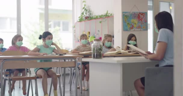 Děti ve škole v maskách sedí ve třídě a poslouchají učitele. Lekce během pandemie ve škole. — Stock video