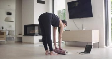 Spor giyimli bir kadın, bilgisayar laptopunda yoga eğitimi alırken esneme egzersizleri yapıyor. Arka planda kanepe ile oturma odasında egzersiz yapan sağlıklı bir kız...