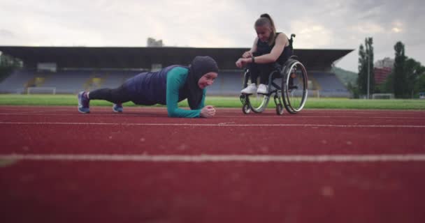 陸上競技場で車いすでウォーミングアップする障害のある女性とヒジャーブのスポーツウェアを着たムスリム女性 — ストック動画