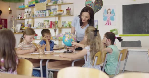 Učitelka s dětmi na hodině zeměpisu, dívající se na zeměkouli. Boční pohled na skupinu různorodých šťastných školáků s glóbus ve třídě ve škole. — Stock video