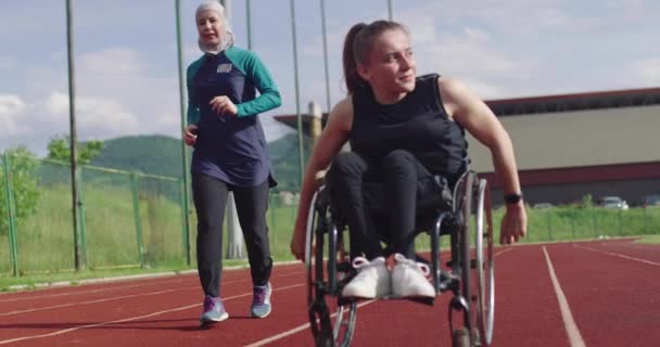 Kobieta z niepełnosprawnością na wózku inwalidzkim na torze treningowym z muzułmanką ubraną w strój sportowy hidżab — Wideo stockowe
