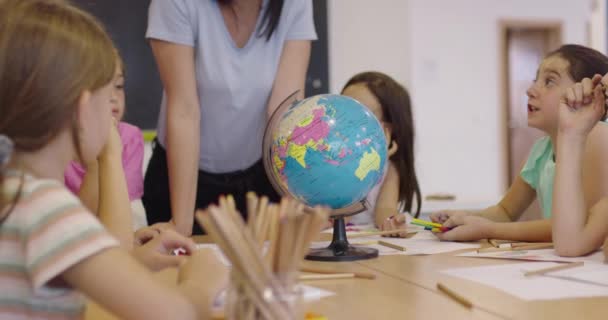 Kvinnelig lærer med barn i geografi som ser på kloden. Sett fra siden av gruppe av diverse glade skolebarn med globus i klasserommet på skolen. – stockvideo