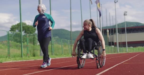 Žena s postižením jezdí na invalidním vozíku na atletické tréninkové dráze s muslimskou ženou na sobě hidžáb sportovní oblečení — Stock video