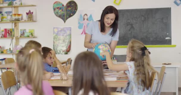 地理学のクラスの子供を持つ女性教師は、地球を見て。学校の教室で地球と多様な幸せな学校の子供たちのグループの側面図. — ストック動画