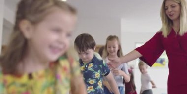 Küçük anaokulu öğrencileri ve bayan öğretmenleri sınıfın içinde egzersiz yapıyorlar. Hula hoop çemberinin üzerinden zıplarken yerde.