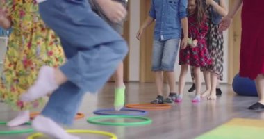 Küçük anaokulu öğrencileri ve bayan öğretmenleri sınıfın içinde egzersiz yapıyorlar. Hula hoop çemberinin üzerinden zıplarken yerde.