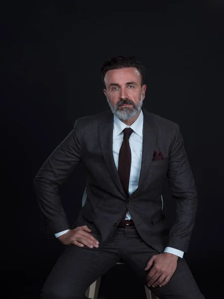 Retrato de un hombre de negocios adulto con traje de moda y sentado en un estudio moderno en una silla elegante contra el fondo negro — Foto de Stock