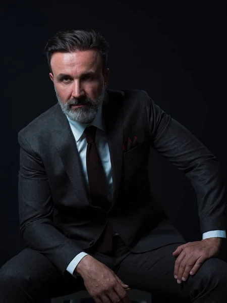 Портрет взрослого бизнесмена в модном костюме и сидящего в современной студии на стильном стуле на черном фоне — стоковое фото