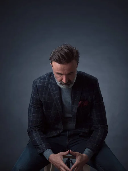 Retrato de empresário adulto vestindo terno moderno e sentado em estúdio moderno em cadeira elegante contra o fundo preto — Fotografia de Stock