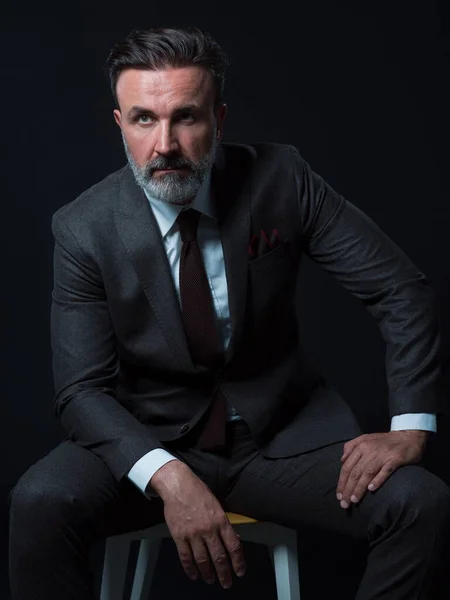 Portré felnőtt üzletember visel trendi öltöny és ül a modern stúdióban stílusos szék ellen fekete háttér — Stock Fotó