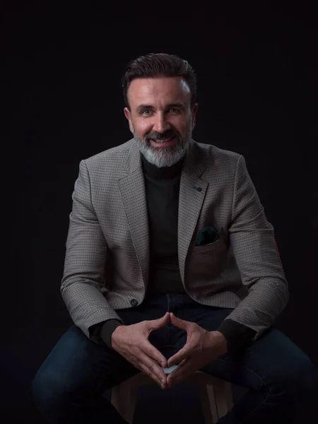 Retrato de un hombre de negocios adulto con traje de moda y sentado en un estudio moderno en una silla elegante contra el fondo negro — Foto de Stock