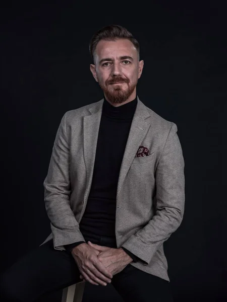 Portrait of adult businessman wearing trendy suit and sitting in modern studio on stylish chair against the black background — Stock Photo, Image