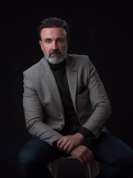 Retrato de un hombre de negocios adulto con traje de moda y sentado en un estudio moderno en una silla elegante contra el fondo negro — Foto de Stock