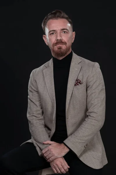 Portret dorosłego biznesmena ubranego w modny garnitur i siedzącego w nowoczesnym studio na stylowym krześle na czarnym tle — Zdjęcie stockowe