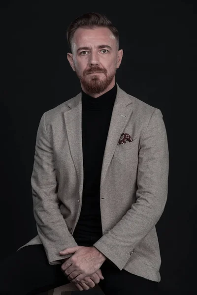 Portret dorosłego biznesmena ubranego w modny garnitur i siedzącego w nowoczesnym studio na stylowym krześle na czarnym tle — Zdjęcie stockowe