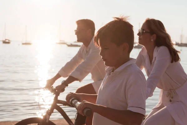Szczęśliwa rodzina ciesząca się pięknym porankiem nad morzem, rodzice jeżdżący na rowerze i ich syn jeżdżący na skuterze elektrycznym. Skupienie selektywne — Zdjęcie stockowe
