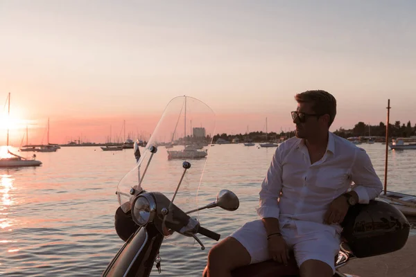 Ein moderner Geschäftsmann in lässiger Kleidung sitzt auf einem Motorroller am Meer und genießt den Sonnenaufgang. Selektiver Fokus — Stockfoto