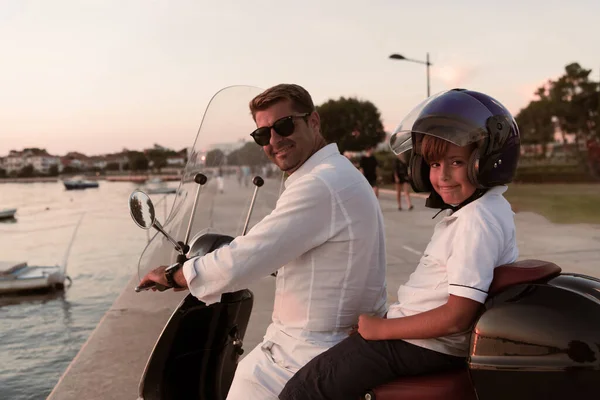 Um pai com seu filho desfruta de umas férias montando uma scooter junto ao mar no início da manhã. Foco seletivo — Fotografia de Stock