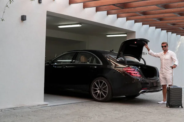 Un hombre mayor en un garaje con gafas y ropa casual moderna saca sus maletas del coche. Enfoque selectivo —  Fotos de Stock