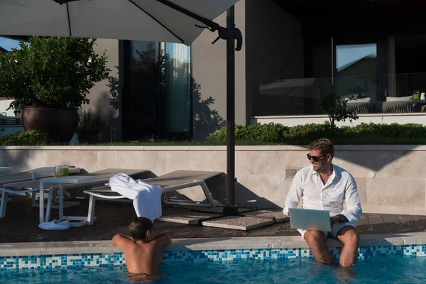 Сучасний літній чоловік насолоджується басейном, працюючи на ноутбуці поруч із сучасним розкішним будинком. Вибірковий фокус — стокове фото