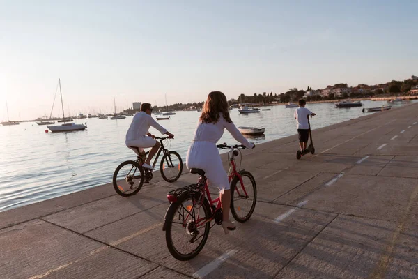 Lycklig familj njuter av en vacker morgon vid havet tillsammans, föräldrar rider en cykel och deras son rider en elektrisk skoter. Selektiv inriktning — Stockfoto