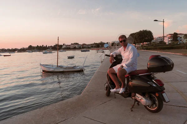 Ένας σύγχρονος ανώτερος επιχειρηματίας με casual ρούχα κάθεται σε ένα σκούτερ δίπλα στη θάλασσα και απολαμβάνει την ανατολή του ηλίου. Επιλεκτική εστίαση — Φωτογραφία Αρχείου