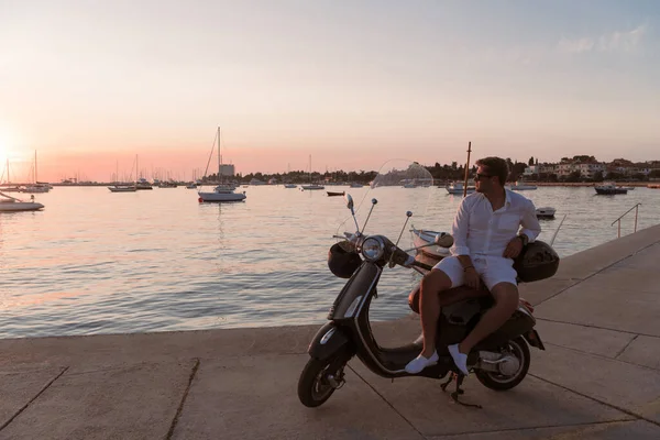 Ein moderner Geschäftsmann in lässiger Kleidung sitzt auf einem Motorroller am Meer und genießt den Sonnenaufgang. Selektiver Fokus — Stockfoto