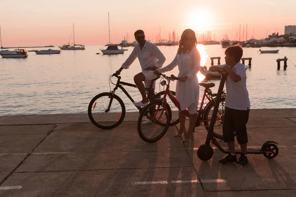 Família feliz desfrutando de uma bela manhã junto ao mar, pais andando de bicicleta e seu filho montando uma scooter elétrica. Foco seletivo — Fotografia de Stock