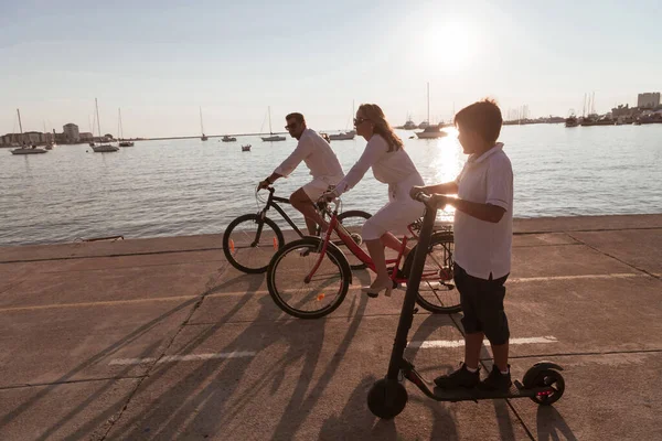 Lycklig familj njuter av en vacker morgon vid havet tillsammans, föräldrar rider en cykel och deras son rider en elektrisk skoter. Selektiv inriktning — Stockfoto