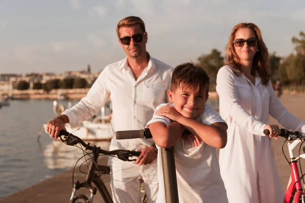 Família feliz desfrutando de uma bela manhã junto ao mar, pais andando de bicicleta e seu filho montando uma scooter elétrica. Foco seletivo — Fotografia de Stock