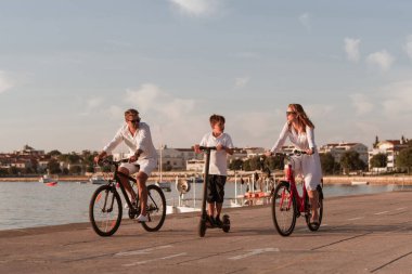 Mutlu bir aile deniz kenarında güzel bir sabah geçiriyorlar, ebeveynler bisiklete biniyor ve oğulları elektrikli scooter kullanıyor. Seçici odak 