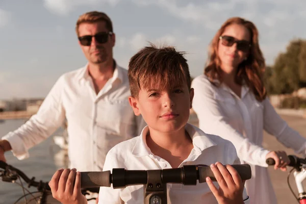 Familia feliz disfrutando de una hermosa mañana junto al mar juntos, los padres montando una bicicleta y su hijo montando un scooter eléctrico. Enfoque selectivo —  Fotos de Stock