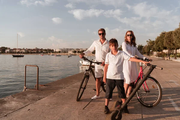 Família feliz desfrutando de uma bela manhã junto ao mar, pais andando de bicicleta e seu filho montando uma scooter elétrica. Foco seletivo — Fotografia de Stock
