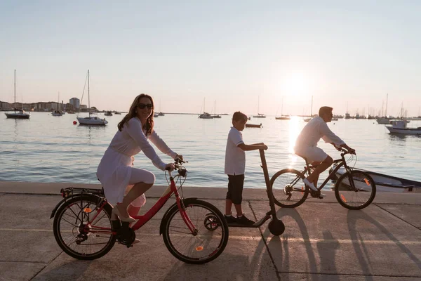 Familia feliz disfrutando de una hermosa mañana junto al mar juntos, los padres montando una bicicleta y su hijo montando un scooter eléctrico. Enfoque selectivo — Foto de Stock