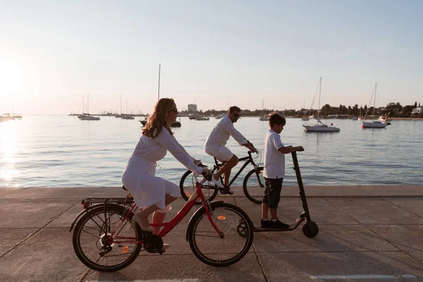 Счастливая семья, наслаждающаяся прекрасным утром у моря вместе, родители катаются на велосипеде, а их сын на электрическом скутере. Селективный фокус — стоковое фото