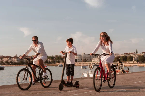 Família feliz desfrutando de uma bela manhã junto ao mar, pais andando de bicicleta e seu filho montando uma scooter elétrica. Foco seletivo — Fotografia de Stock